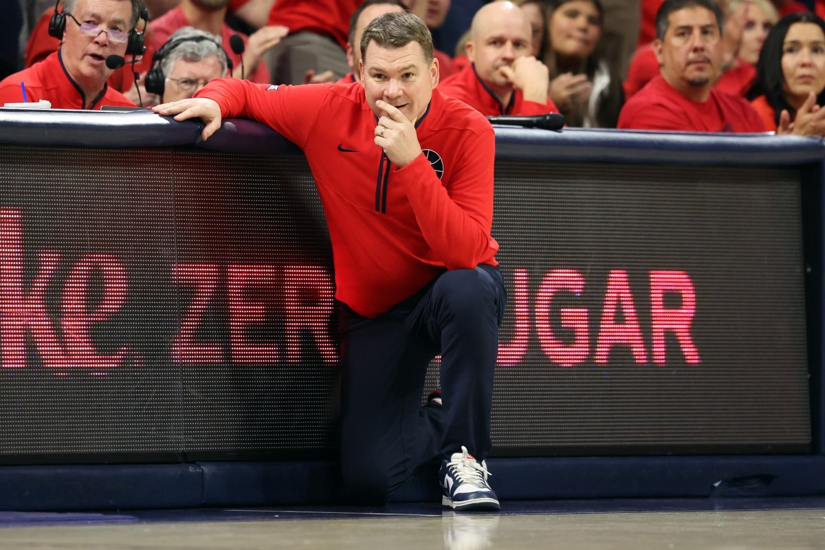 Arizona men's basketball coach Tommy Lloyd crouches on the sideline with his head resting on his hand and his other arm on the scorer's table