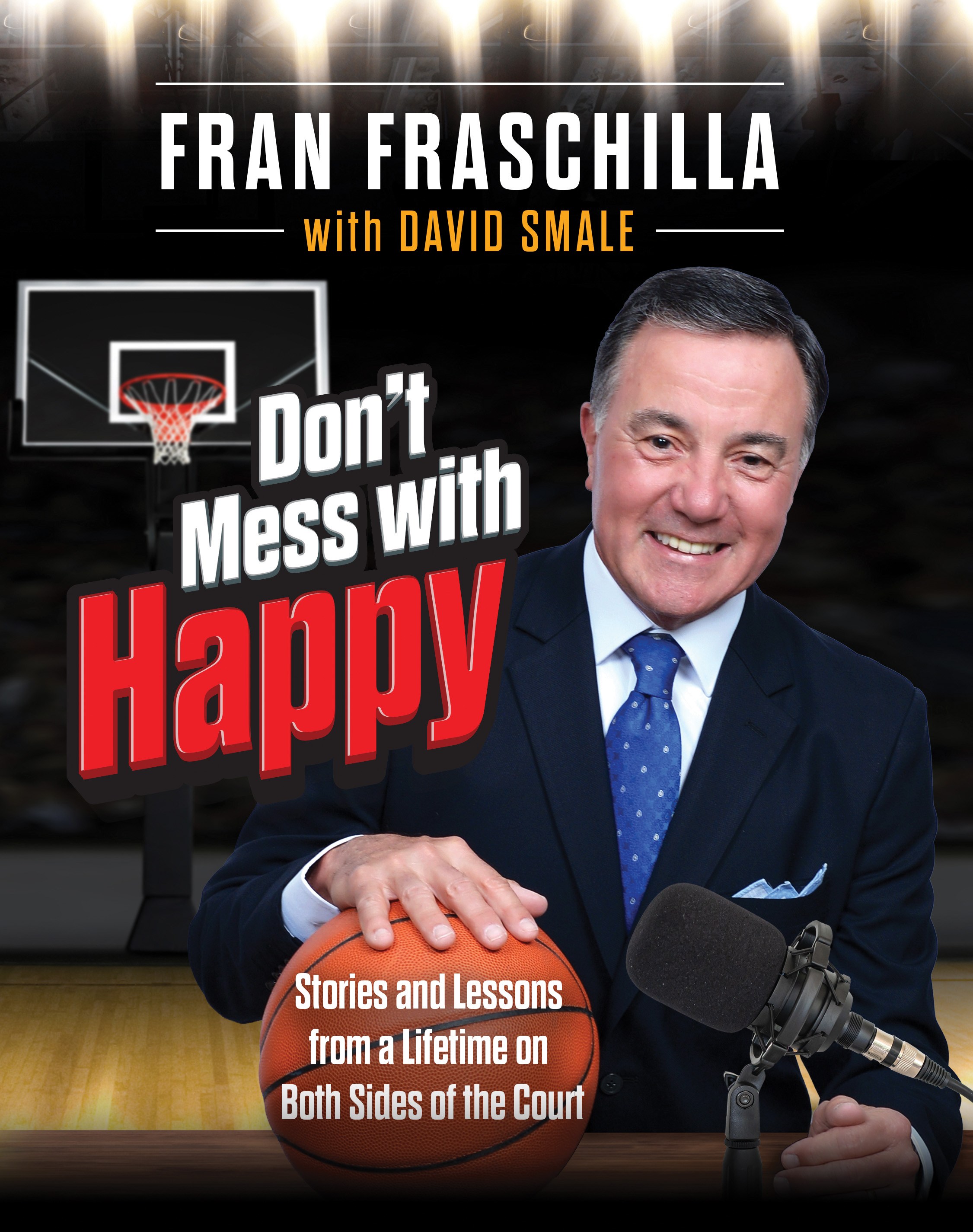 Fran Fraschilla smiling, hand on a basketball, next to a microphone, with a basketball hoop in the background.