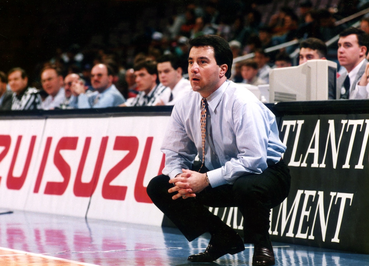 A man in a light blue shirt and patterned tie crouches intently on a basketball court, watching the game.