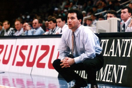 A man in a light blue shirt and patterned tie crouches intently on a basketball court, watching the game.