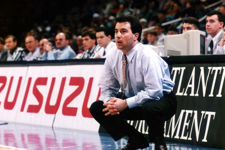 A man in a light blue shirt and patterned tie crouches intently on a basketball court, watching the game.