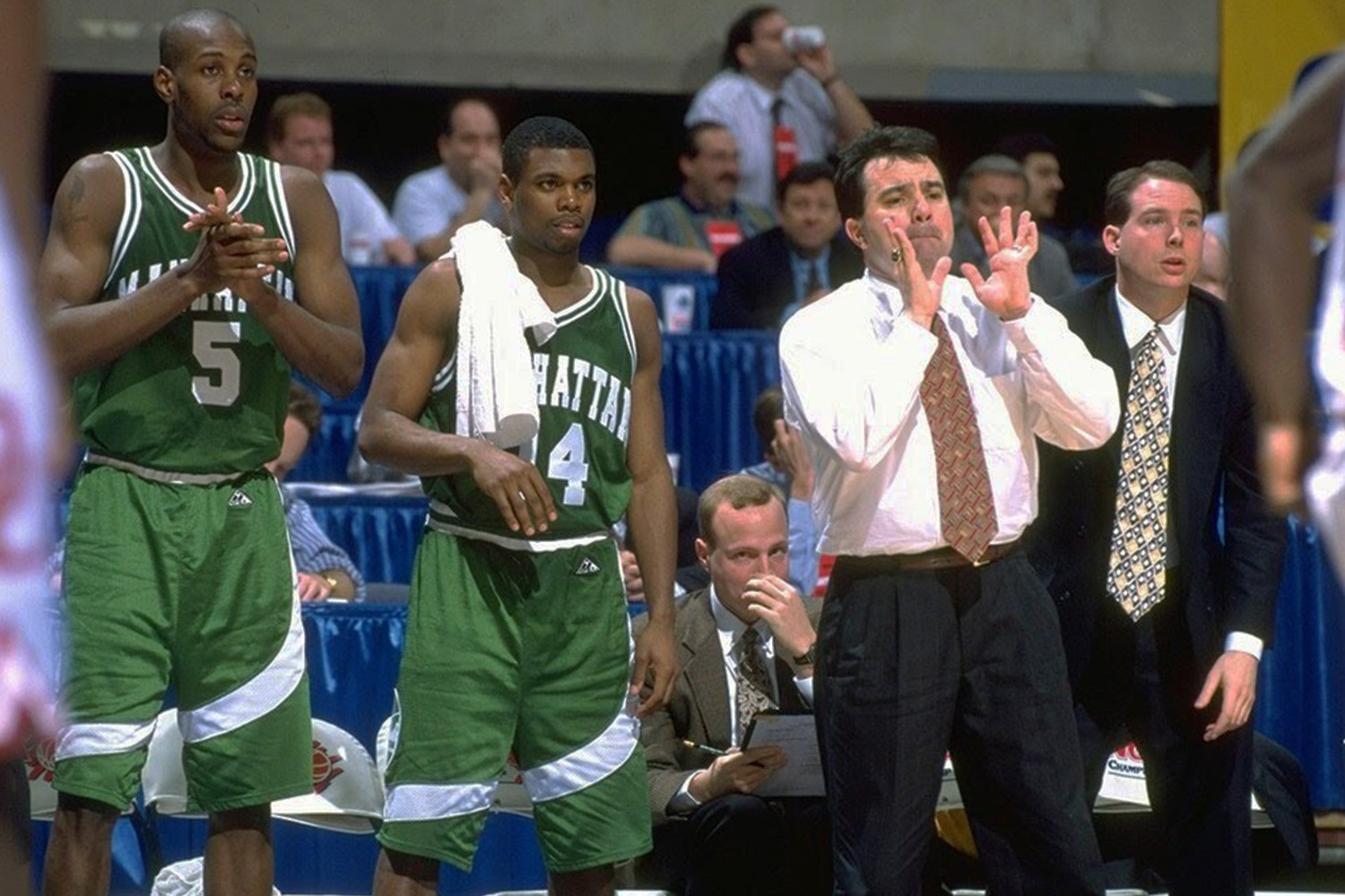 Two basketball players in green jerseys stand next to a coach in a white shirt, gesturing with his hands.